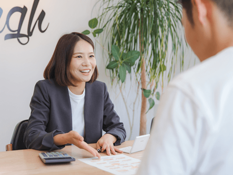 笑顔で説明する女性スタッフと向かい合って話を聞いている男性。テーブルには資料と電卓が置かれている。